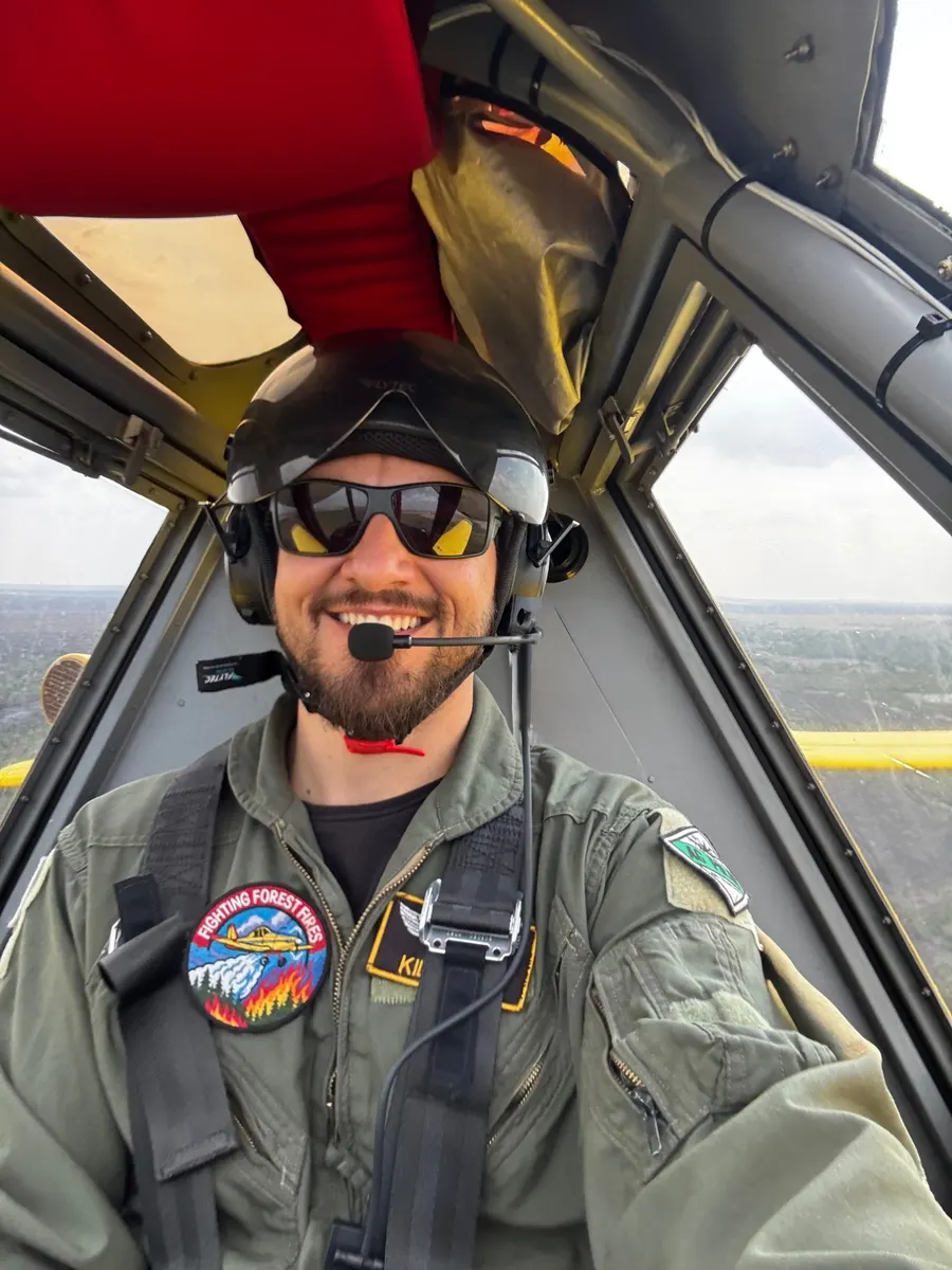 Guilherme Kilian na cabine do avião em voo, de capacete e macacão com os patches de combate a incêndio